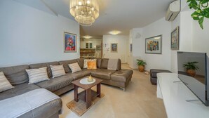 Un soggiorno moderno con un divano componibile, un tavolino da caffè, una TV e un lampadario. Si intravede una cucina sullo sfondo.