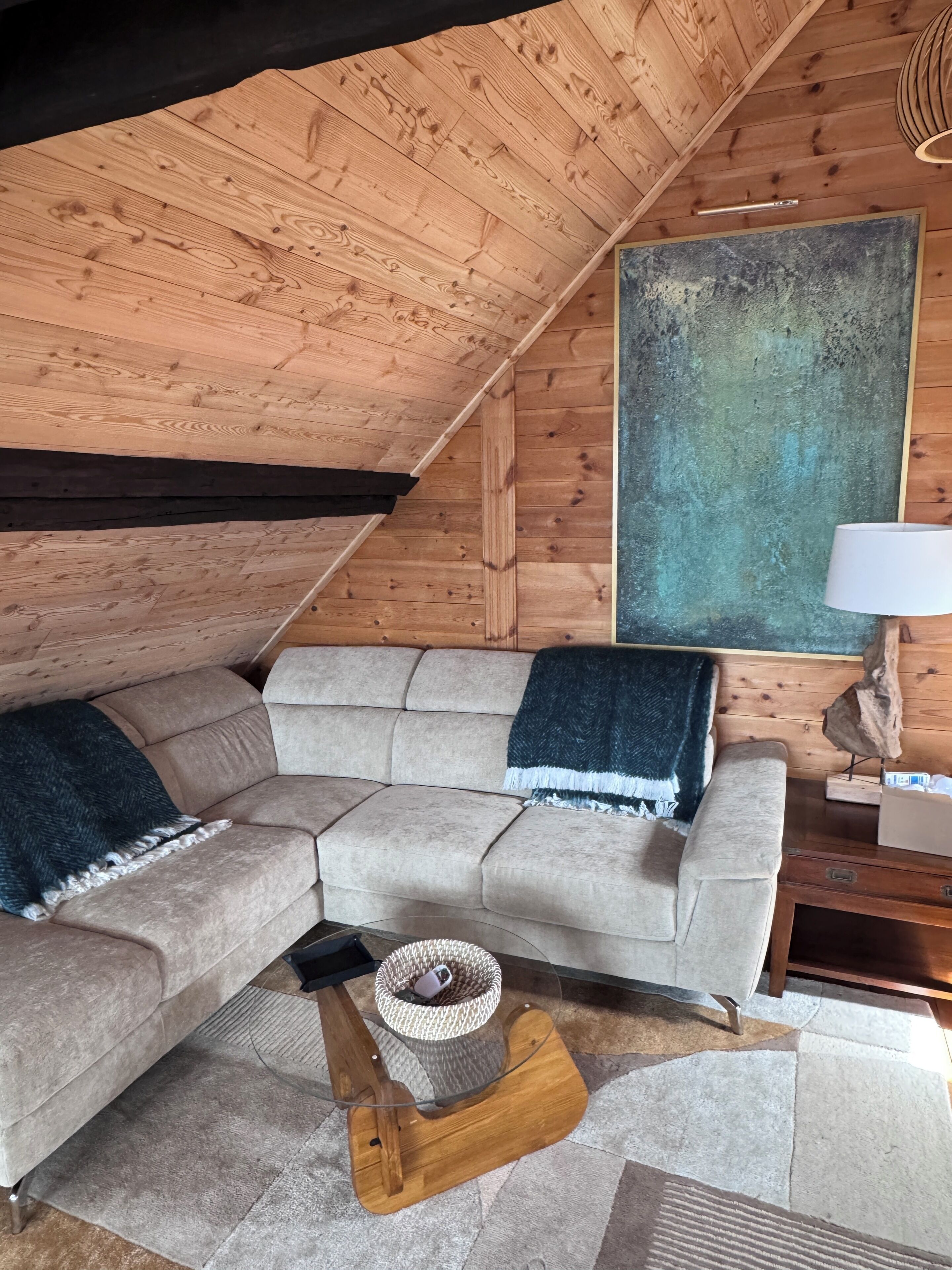 A cozy loft bedroom with a sloped ceiling, a sectional sofa, a large abstract painting, and a bedside table with a lamp.