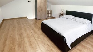Une chambre avec un plancher en bois, un lit recouvert d’une courtepointe blanche, une tête de lit sombre, une armoire en bois et une table de chevet avec une lampe.