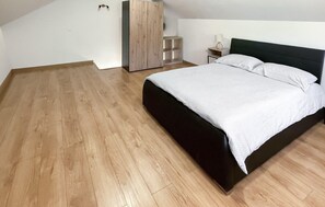Un dormitorio con suelo de madera, una cama con colcha blanca, cabecero oscuro, armario de madera y mesita de noche con lámpara.