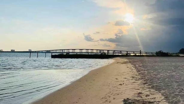 Una spiaggia con un molo che si protende in acque calme, sotto un cielo parzialmente nuvoloso con il sole basso sull'orizzonte.