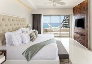 A modern hotel room with a large bed, a television mounted on the wall, and a view of the sea through a glass door.