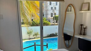 A hotel room with a bed, a desk, a chair, a large window with a view of a swimming pool and palm trees. - Comfortable and spacious room w/alberca (Cancún)