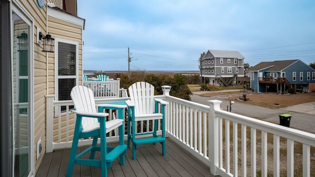 Una terraza con dos sillas Adirondack blancas y una mesa, con vistas a un área residencial con casas y un cielo despejado.