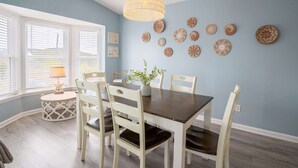 A dining area with a dark wooden table, white chairs, a hanging light fixture, and wall decor. - So Free - Dogs Allowed - Walk to Beach (North Topsail Beach)