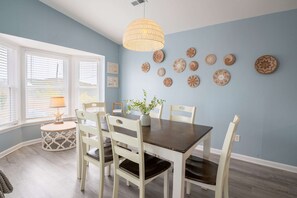 A dining area with a dark wooden table, white chairs, a hanging light fixture, and wall decor. - So Free - Dogs Allowed - Walk to Beach (North Topsail Beach)