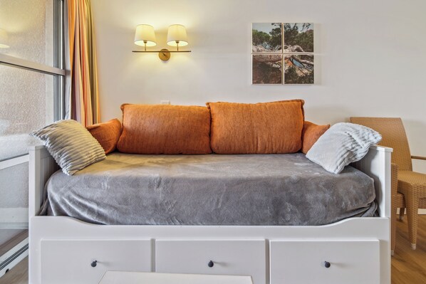 Une chambre à coucher moderne avec un canapé gris, deux coussins, une table de chevet blanche avec des tiroirs et une lampe fixée au mur.