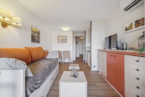 Ein modernes, helles Zimmer mit einer Couch, einem kleinen Tisch mit einem Laptop, einer Küchenzeile mit Schränken und einem Fernseher.