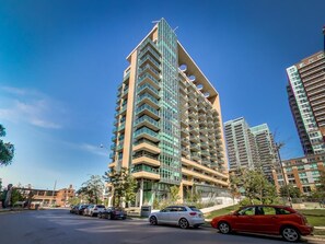 A high-rise building with multiple balconies, a parking lot with cars, and a clear blue sky. - Loft in Liberty Village (Steps to Fifa World Cup Stadium) (Toronto)