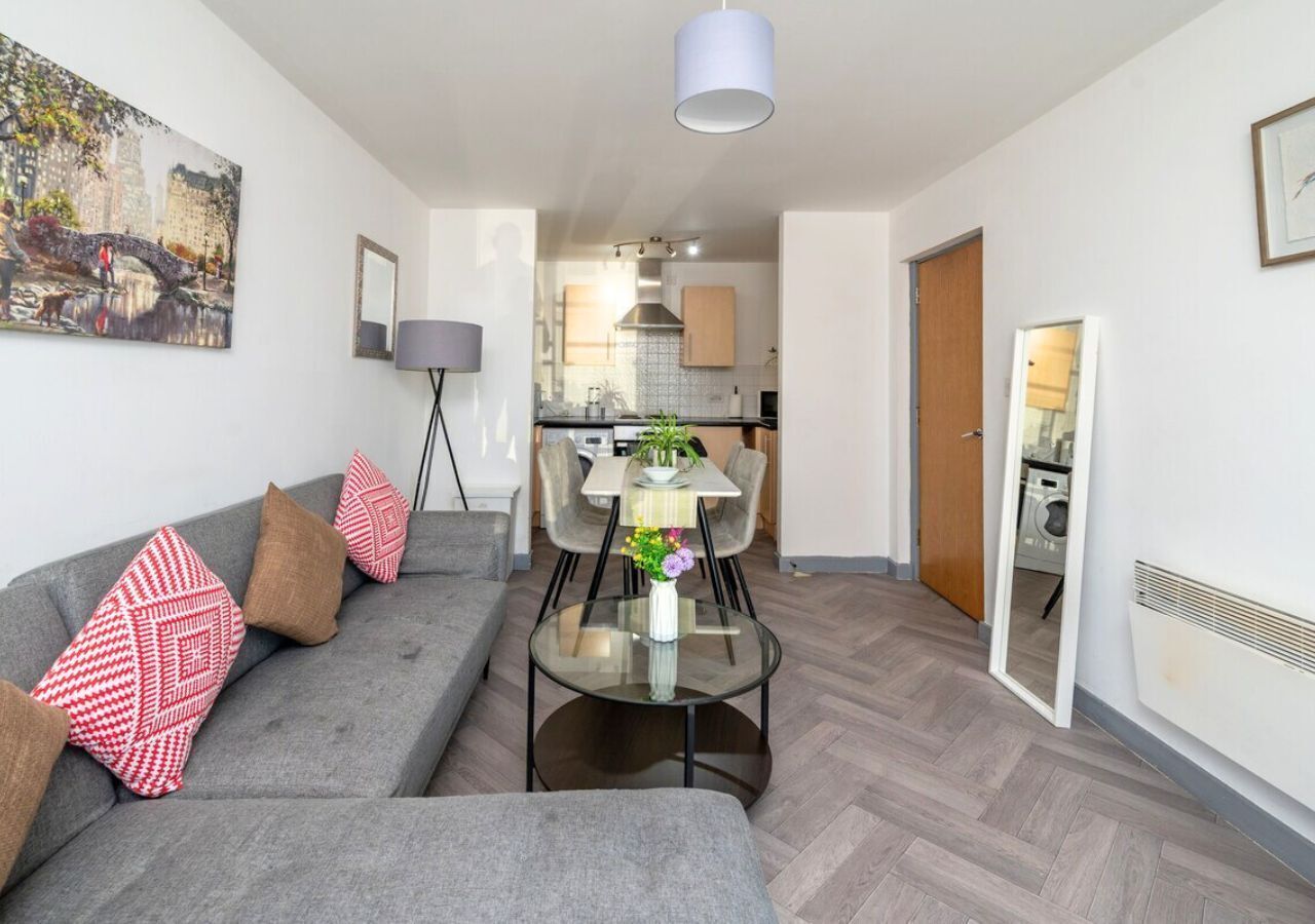 Een moderne woonkamer met een grijze bank, een glazen salontafel en een eethoek met een witte tafel en stoelen.