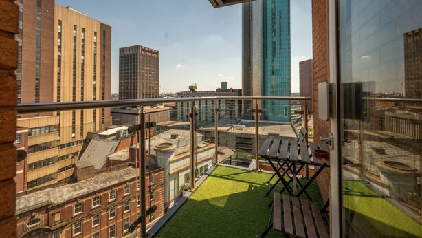 Un balcón con vista a edificios circundantes, una mesa de madera y una zona ajardinada.