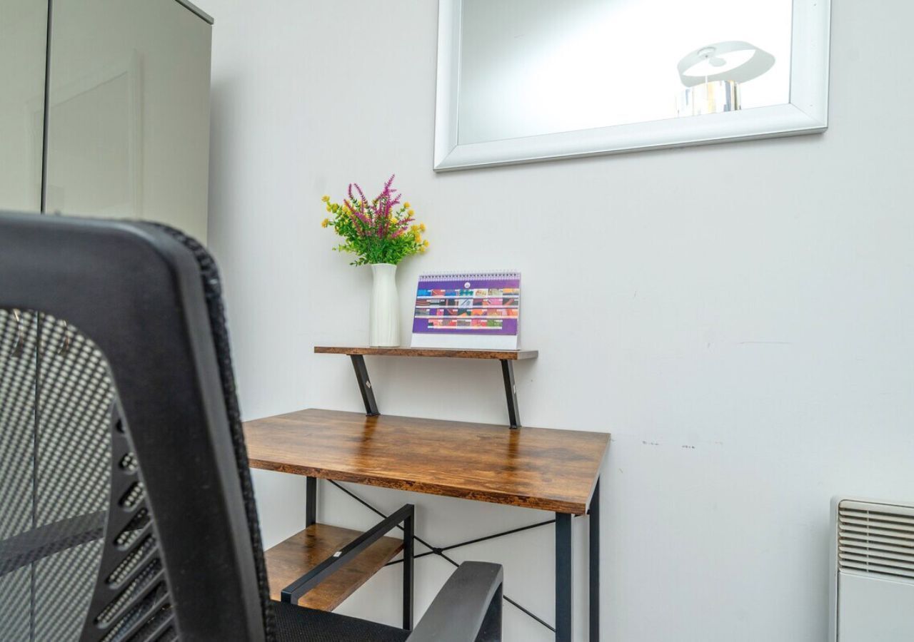 Ein kleiner Holztisch mit einer weißen Vase voller Blumen und einem bunten Buch, vor einer weißen Wand.