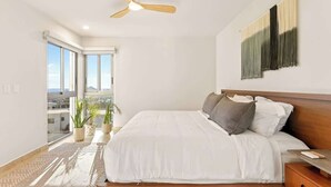 Une chambre à coucher moderne, dotée d’un grand lit, d’un ventilateur de plafond et offrant une vue sur la ville grâce à de grandes fenêtres.
