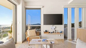 Un salon moderne doté d’une grande fenêtre, d’un téléviseur à écran plat fixé au mur et d’un coin salon confortable avec un canapé et un fauteuil.