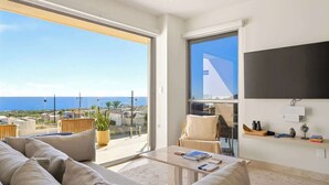 Un salon moderne avec un grand canapé, une table basse et un téléviseur fixé au mur. Une grande fenêtre offre une vue sur l’océan, et un balcon est aménagé avec du mobilier d’extérieur.