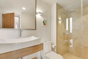 Een moderne badkamer met een gebogen witte wastafel, een houten opbergmeubel, een toilet en een douchecabine met glazen wanden.