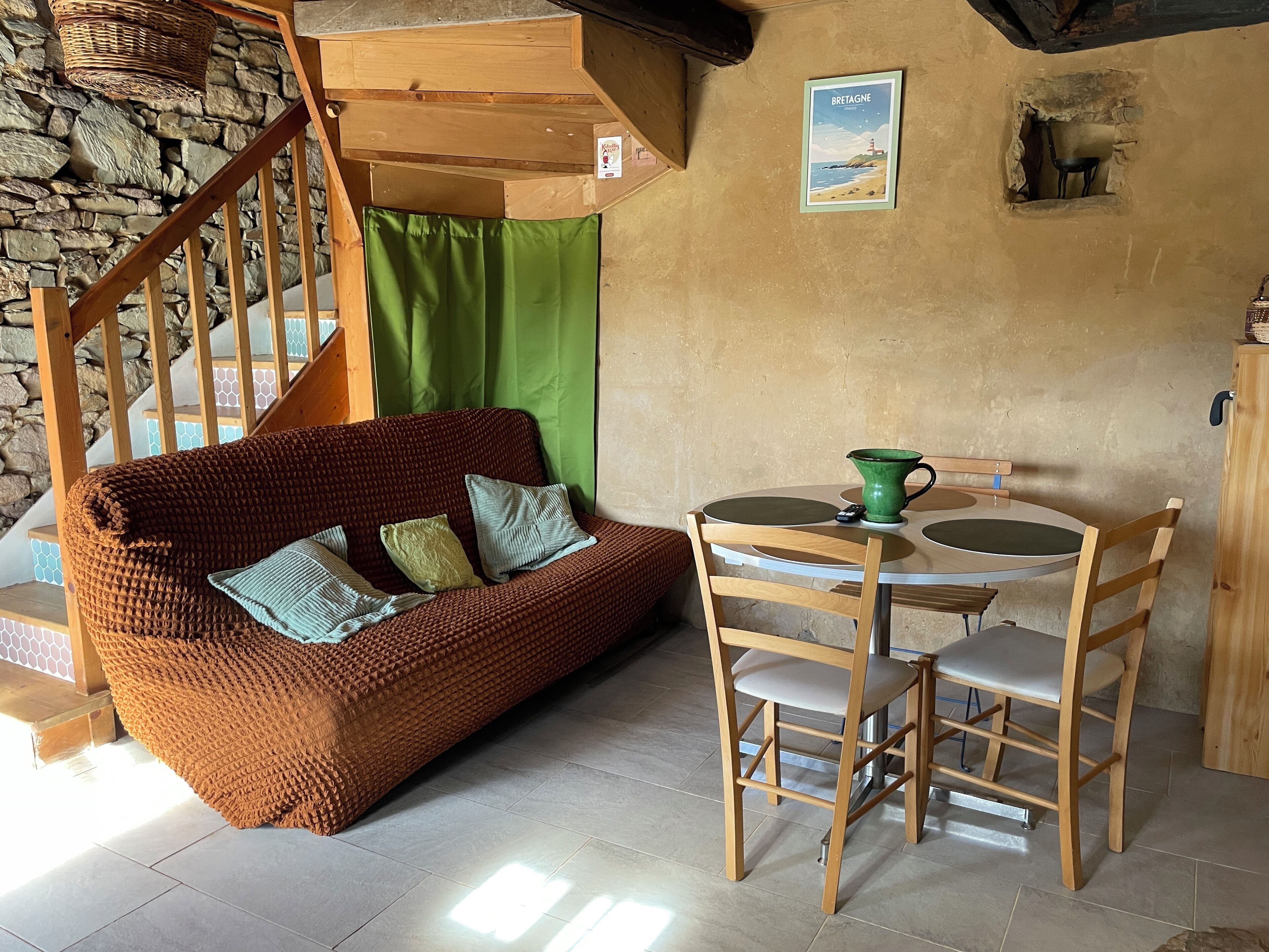 A cozy room with a wooden staircase, a woven sofa, a green curtain, a small table with a green pitcher, and a wall-mounted picture of Brittany.