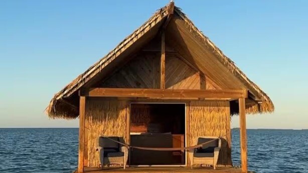 Una casa flotante de madera con techo de paja, dos sillas y una tabla de surf sobre el agua.