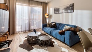 Uma sala de estar moderna com um sofá azul, duas mesas de centro redondas, um laptop em uma das mesas e uma janela grande com cortinas leves.
