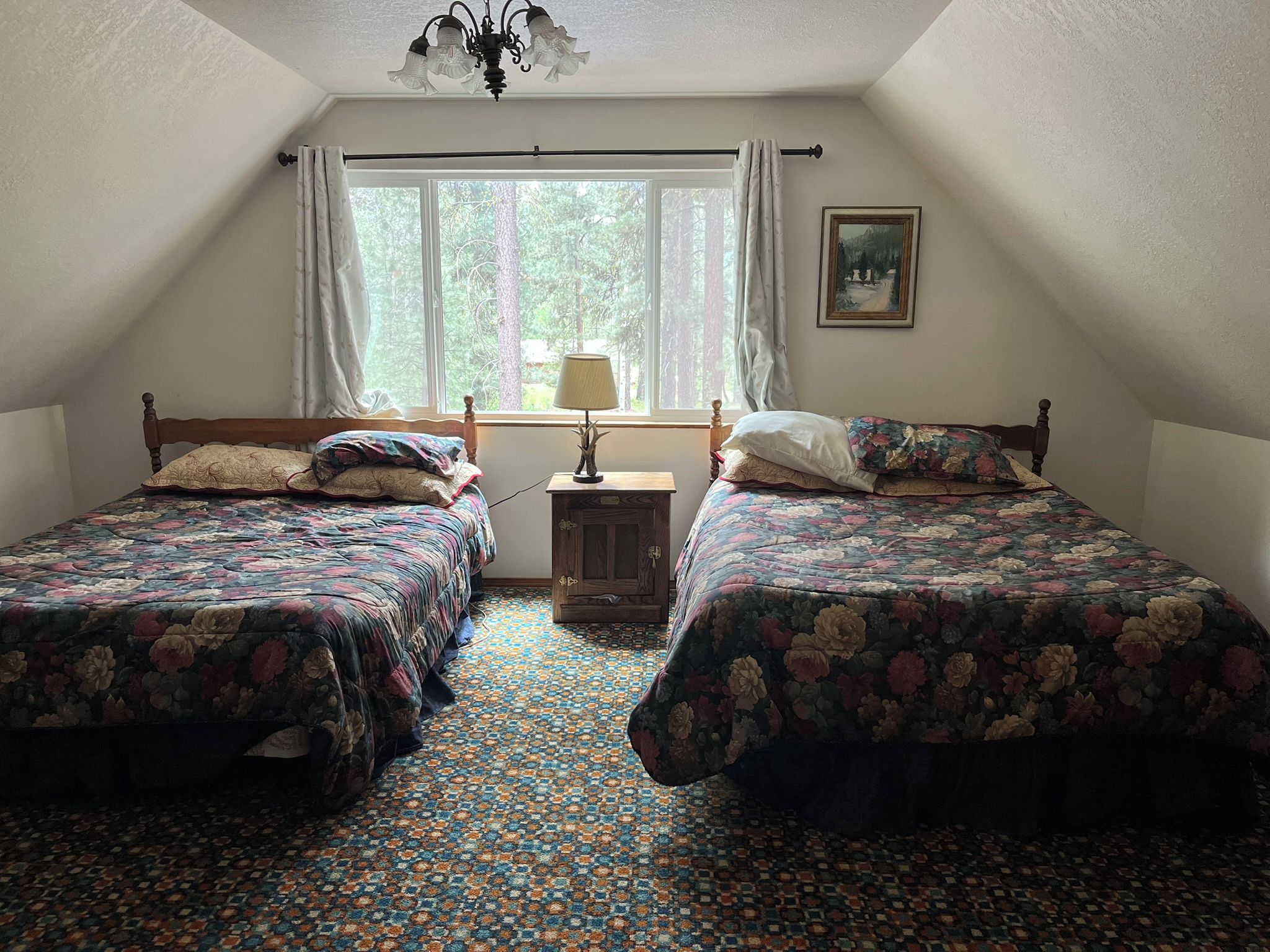 A bedroom with two beds, a window with curtains, a lamp, and a painting on the wall.