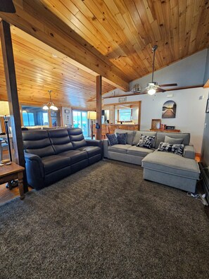 A spacious living room with a wooden ceiling, a black leather sofa, a grey sectional sofa, and a ceiling fan. - Savor a Sunset on Waneta Lake! (Dundee)