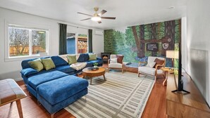 Una sala de estar con un sofá seccional azul, una mesa redonda de centro, un ventilador de techo y un mural de un bosque.