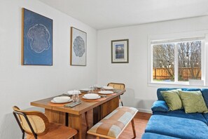Un coin repas avec une table en bois, des chaises et un banc. Des tableaux encadrés ornent le mur, et une fenêtre offre une vue sur les arbres.