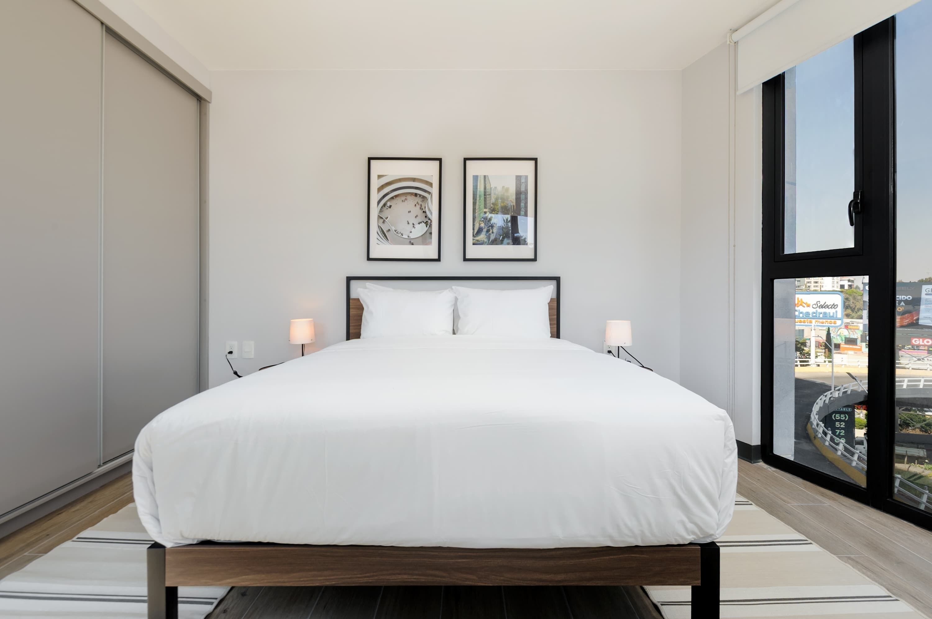 A modern bedroom with a large bed, two framed pictures on the wall, and a view of the city through a large window.