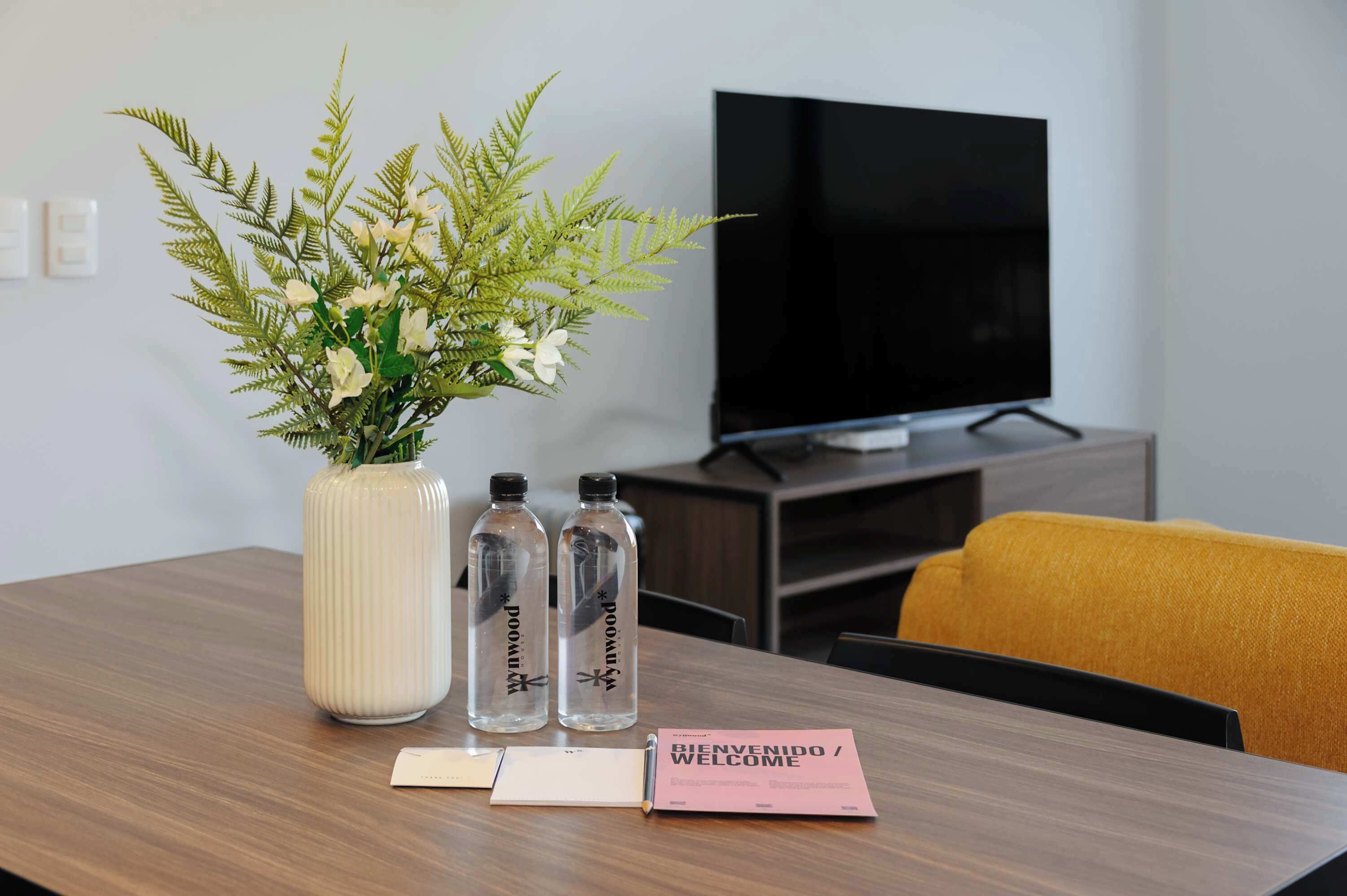 A hotel room with a wooden table, a potted fern, two water bottles, a TV, and a welcome sign.