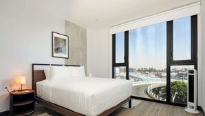 Een moderne hotelkamer met een groot bed, een nachtkastje, een lamp en een uitzicht op een stadsbeeld door grote ramen.