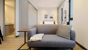 Un salon moderne avec un canapé gris, une petite table et un plaid à motifs. Deux tableaux encadrés sont accrochés au mur, et un miroir se trouve au-dessus du canapé.