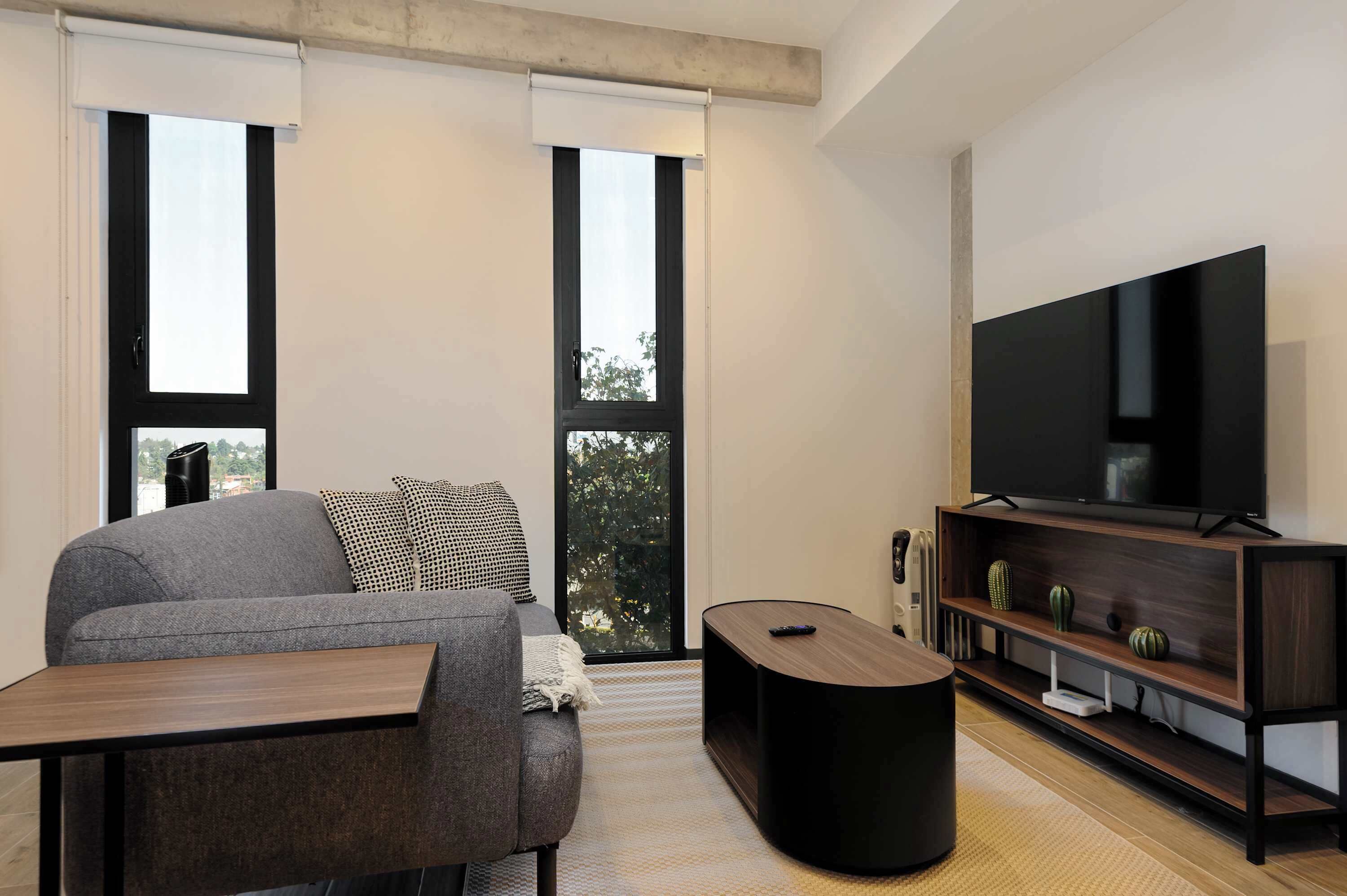 A modern living room with a grey sofa, a wooden coffee table, a flat-screen TV on a wooden stand, and a large window with blinds.