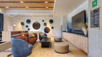 Un salon moderne comprenant un canapé, un fauteuil, un pouf et une table basse. Un téléviseur à écran plat est fixé au mur, et un panneau indique un espace de détente.