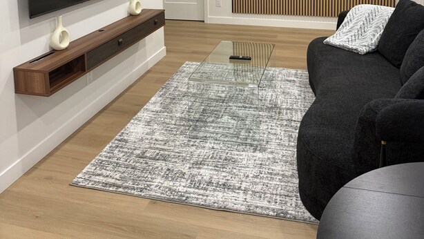 Un salon moderne avec un téléviseur à écran plat fixé au mur, un canapé foncé, une table basse en verre et un tapis à motifs.