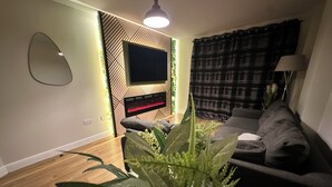 Una sala de estar moderna con un televisor de pantalla plana montado en una pared con estampado geométrico, un sofá gris, una mesa de centro de madera y una planta grande en maceta al frente.