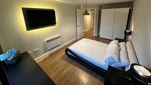 Una habitación de hotel moderna con una cama grande, un televisor de pantalla plana en la pared, una mesita de noche con lámpara y un armario.