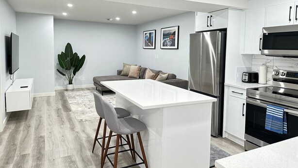 A modern kitchen with a central island, stainless steel appliances, and a grey sofa in the living area. - Modern 2 Bedroom Suite (Airdrie)