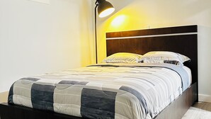 A modern bedroom with a wooden headboard, a quilted bedspread, and a floor lamp providing light. - Modern 2 Bedroom Suite (Airdrie)