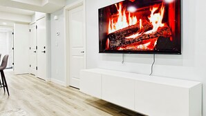 A modern living room with a large flat-screen TV mounted on a white wall, a wooden floor, and a white entertainment unit. - Modern 2 Bedroom Suite (Airdrie)