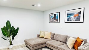 A modern living room with a sectional sofa, a large potted plant, and two framed pictures on the wall. - Modern 2 Bedroom Suite (Airdrie)
