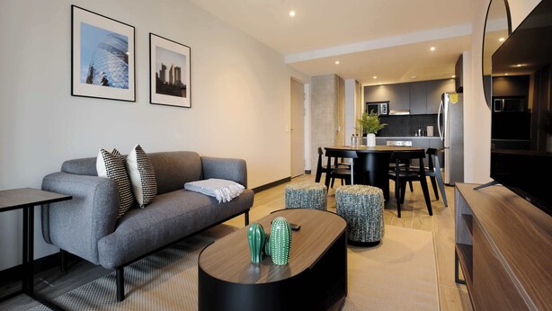 Una sala moderna con un sofá gris, una mesa de centro con objetos decorativos, una zona de comedor con una mesa y sillas, y una cocina con armarios oscuros y un televisor grande.
