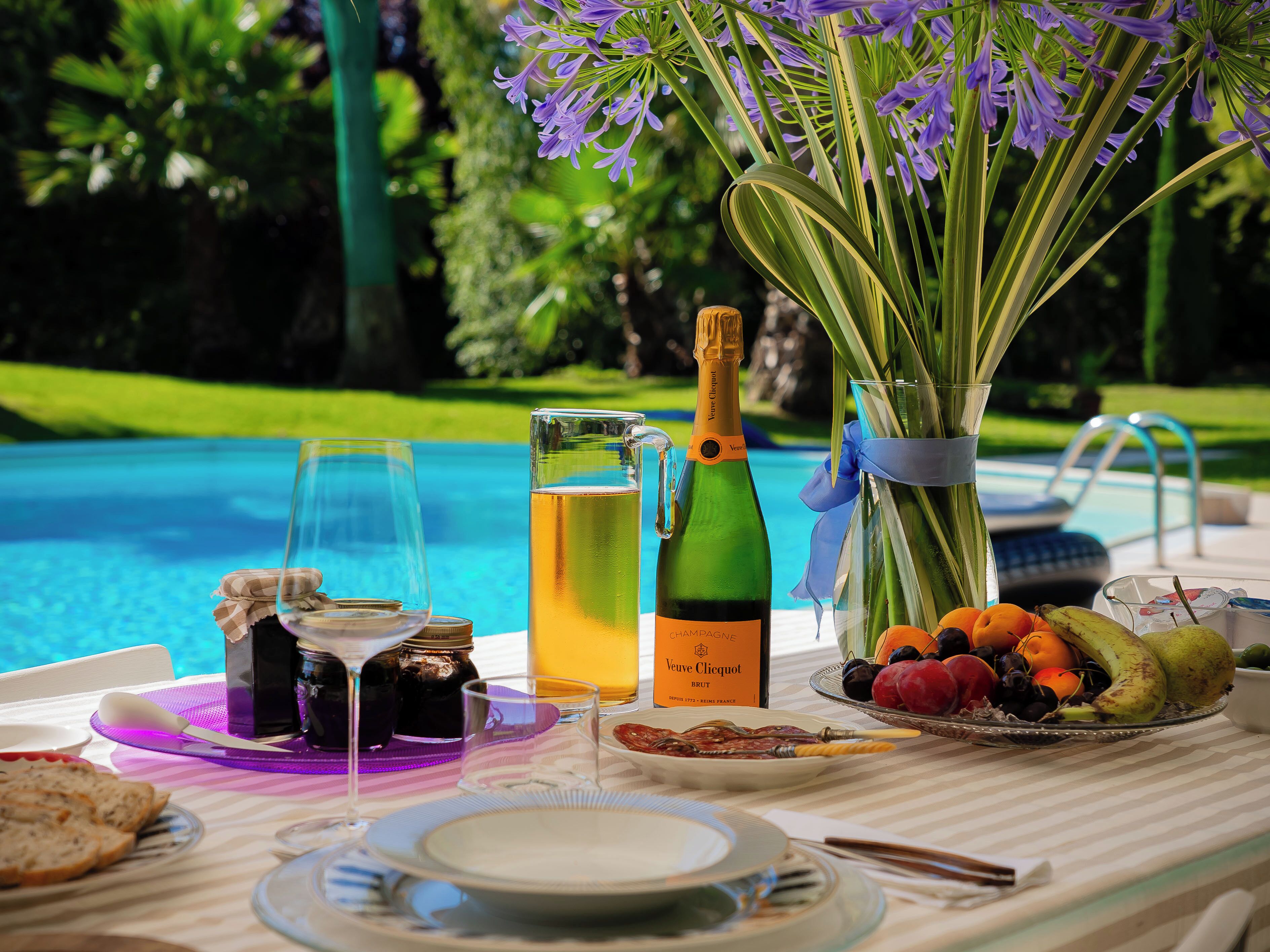 Una mesa dispuesta con una botella de Veuve Clicquot Brut champagne, un vaso de jugo de naranja, una bandeja de fruta y un florero junto a una piscina.