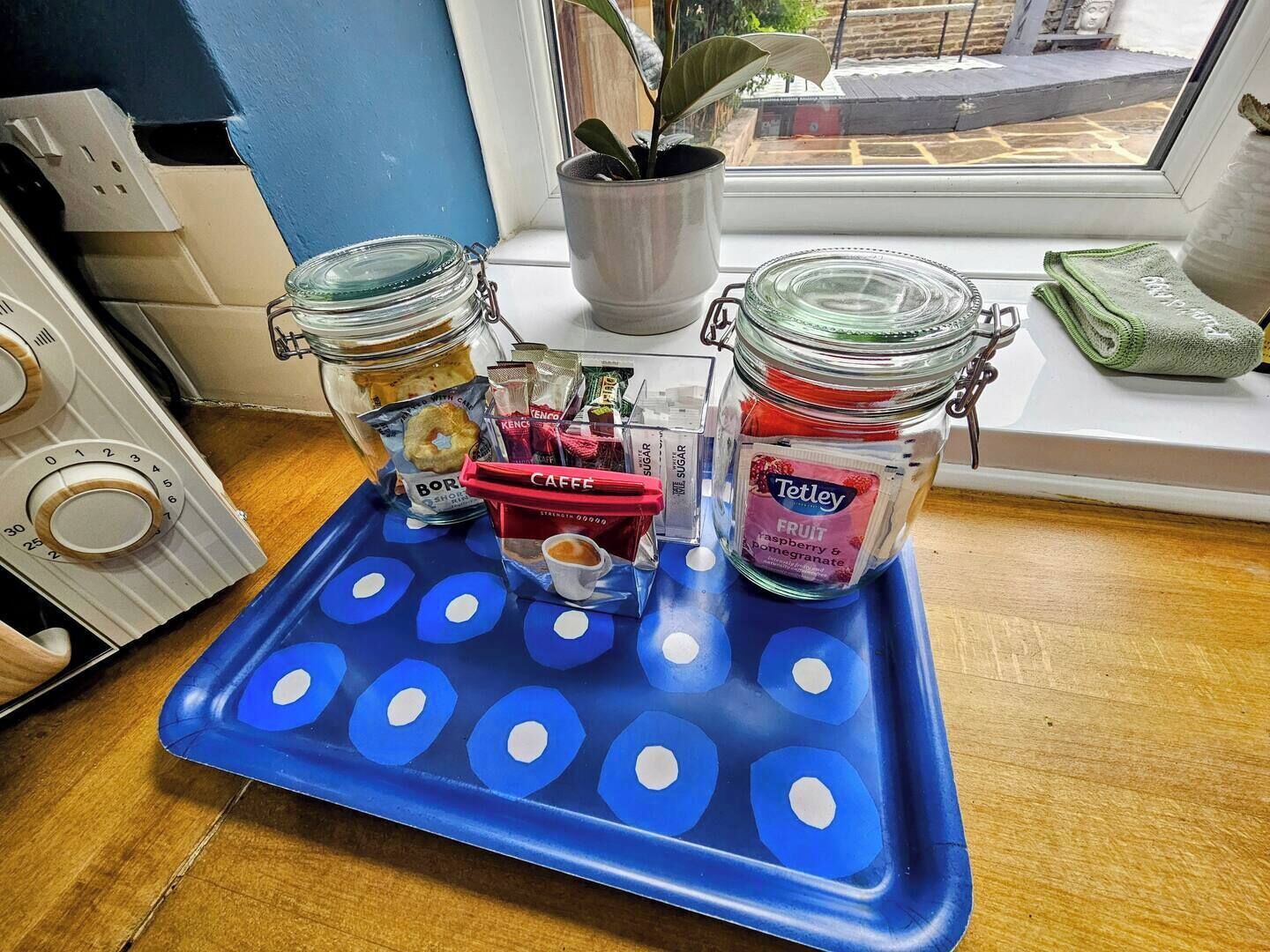 A kitchen counter with a blue tray holding jars of coffee and tea, a kettle, and a plant.