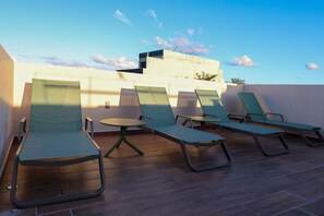 Eine Dachterrasse mit vier Liegestühlen und einem kleinen runden Tisch, gestaltet im modernen Architekturstil mit flachem Dach und einer weißen Wand im Hintergrund.