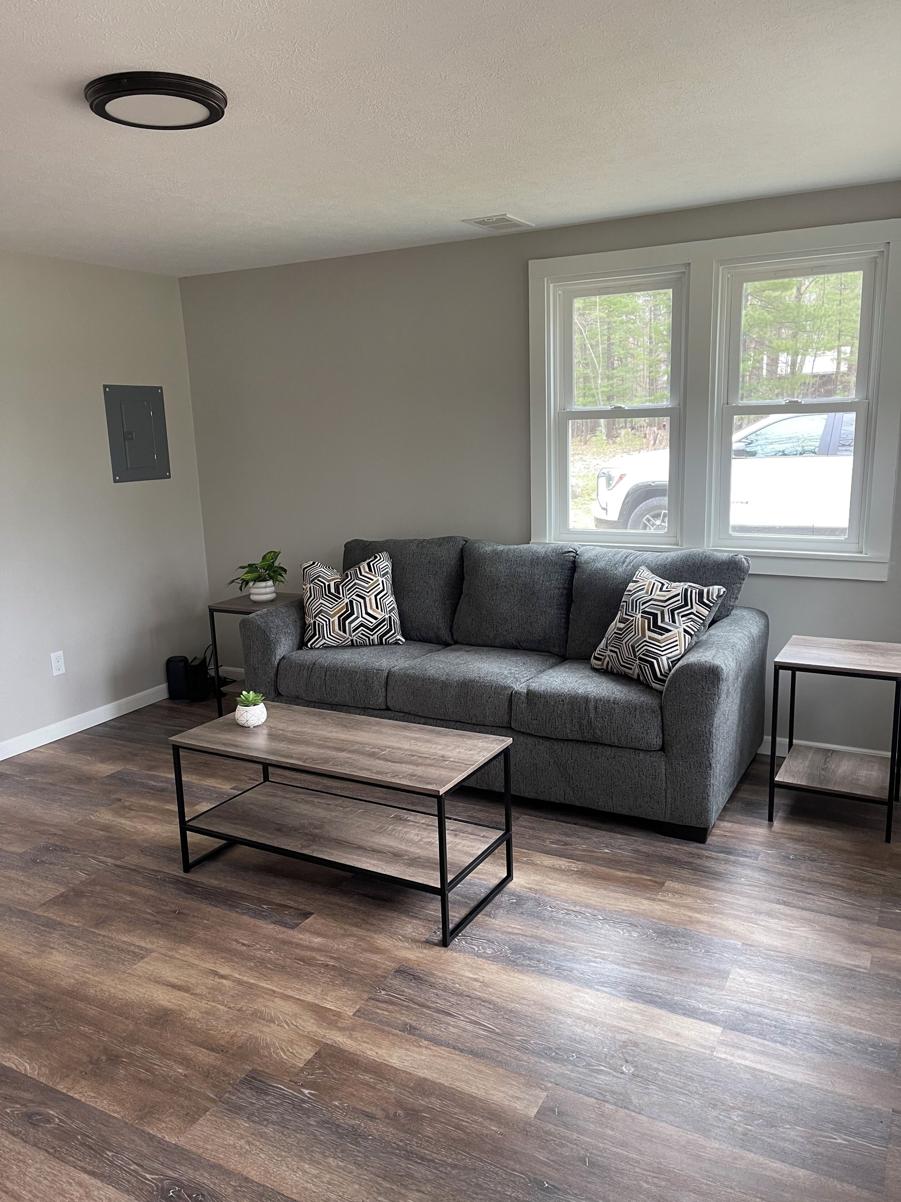 A living room with a grey sofa, patterned cushions, a wooden coffee table, and a window with a view of a parked car.