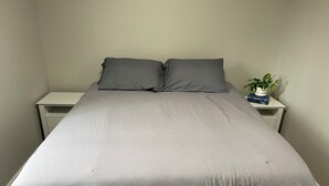 A bed with a grey bedspread, two pillows, a bedside table with a plant, and a book.