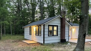 A small, single-story cabin with a brick chimney, white siding, and black trim. It features two windows with black frames and a door with a light fixture.