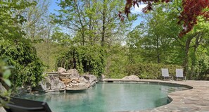 Una piscina con un elemento rocoso natural, rodeada de árboles y un patio con sillas.