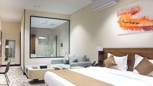 Quarto de hotel com uma cama grande, um sofá, uma mesa de trabalho e um banheiro visível através de um painel de vidro.