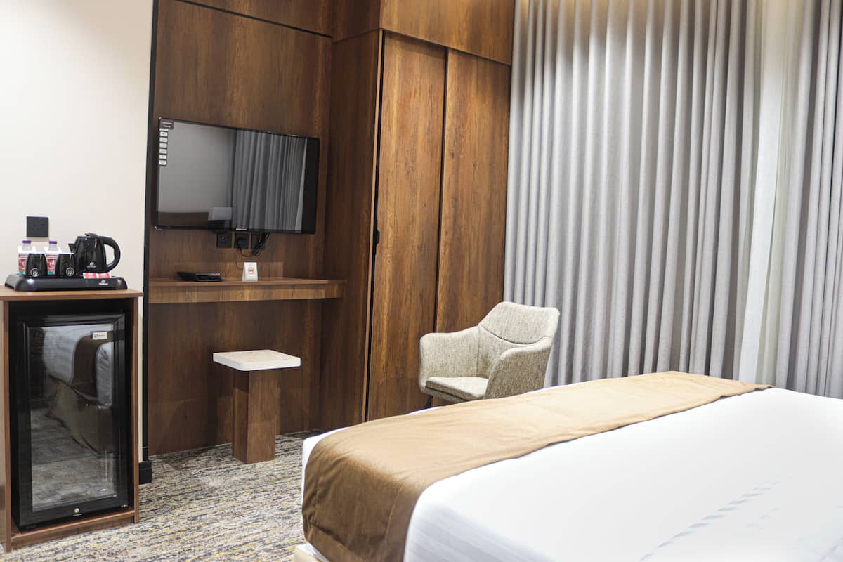 A hotel room with a bed, a desk with a television, a chair, and a minibar.